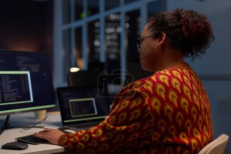 Foto de Vista trasera de la mujer afroamericana sentada en su lugar de trabajo con computadoras, que trabaja en la oficina hasta altas horas de la noche - Imagen libre de derechos