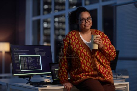 Foto de Retrato del programador afroamericano mirando la cámara mientras trabaja hasta altas horas de la noche en la oficina con una taza de café - Imagen libre de derechos