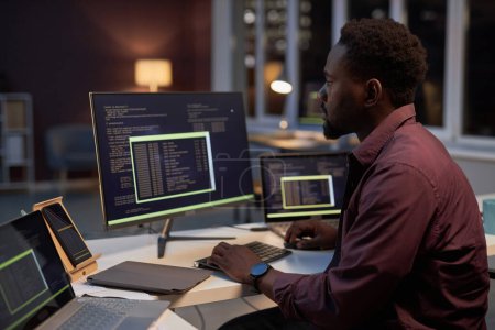Foto de Joven programador afroamericano concentrándose en trabajar con códigos mientras está sentado en la mesa y trabajando en la computadora - Imagen libre de derechos