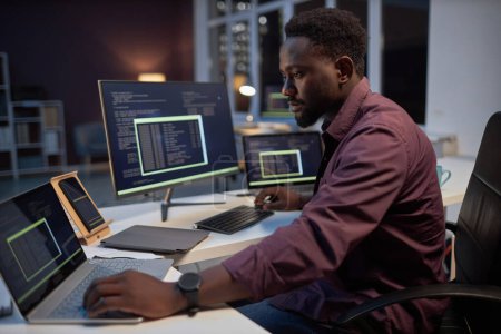 Téléchargez les photos : Programmeur afro-américain se concentrant sur son travail en ligne sur les ordinateurs tout en travaillant dans le bureau jusqu'à tard le soir - en image libre de droit