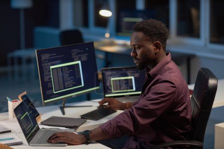 Foto de Desarrollador afroamericano que trabaja con el programa de computadora en la computadora mientras está sentado en su lugar de trabajo en la noche - Imagen libre de derechos