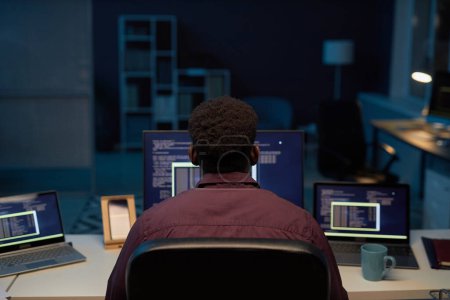 Foto de Vista trasera del programador sentado en su lugar de trabajo frente a las computadoras y trabajando hasta altas horas de la noche - Imagen libre de derechos