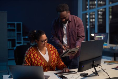 Foto de Hombre mostrando la presentación en la tableta PC a su colega mientras ella trabaja en su lugar de trabajo con la computadora durante el trabajo tardío en la oficina - Imagen libre de derechos