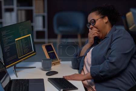 Foto de Programadora cansada bostezando mientras trabaja en su lugar de trabajo en la computadora hasta altas horas de la noche durante la fecha límite - Imagen libre de derechos