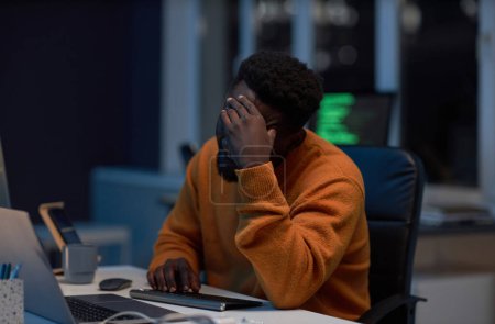 Téléchargez les photos : Afro-américain fatigué développeur faire des heures supplémentaires à son lieu de travail avec ordinateur dans le bureau sombre - en image libre de droit