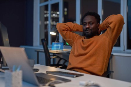 Téléchargez les photos : Développeur afro-américain se reposant sur son lieu de travail après le travail dans un bureau sombre - en image libre de droit