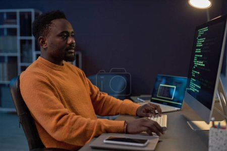 Téléchargez les photos : Développeur afro-américain travaillant avec des codes de sécurité de programme sur ordinateur assis sur son lieu de travail dans le bureau informatique sombre - en image libre de droit