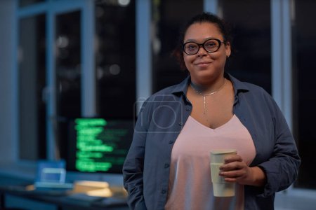 Téléchargez les photos : Portrait de femme afro-américaine en lunettes regardant la caméra tout en se tenant debout avec du café dans le bureau informatique - en image libre de droit