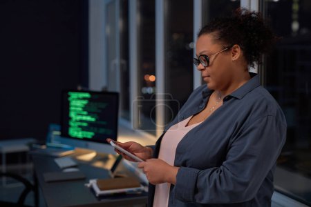 Foto de Mujer leyendo mensaje en su teléfono inteligente mientras trabaja en la oficina hasta altas horas de la noche - Imagen libre de derechos