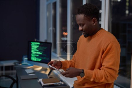 Téléchargez les photos : Homme d'affaires afro-américain lisant un message sur son smartphone tout en travaillant au bureau jusqu'à tard dans la nuit - en image libre de droit