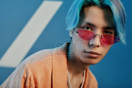 Téléchargez les photos : Portrait de jeune homme avec des lunettes de soleil roses et coiffure bleue regardant la caméra sur fond bleu - en image libre de droit