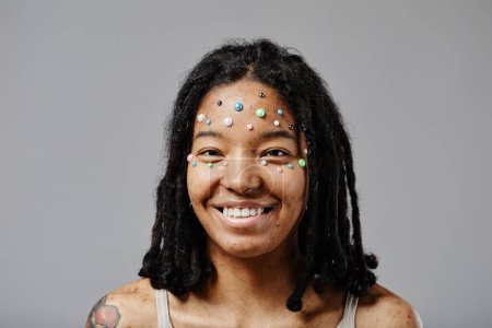 Foto de Mínimo retrato de vista frontal de mujer joven negra sin maquillaje y perlas como decoración de la cara sonriente - Imagen libre de derechos