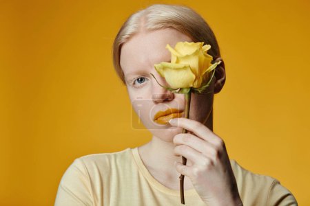 Minimal fashion portrait of young woman with albinism holding rose against contrasting yellow background
