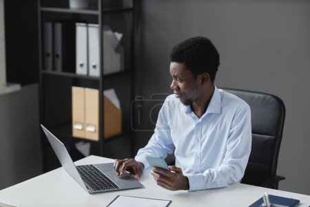 Foto de Retrato de alto ángulo del joven hombre de negocios negro usando el ordenador portátil mientras está sentado en el escritorio del lugar de trabajo en la oficina, espacio de copia - Imagen libre de derechos