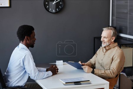 Téléchargez les photos : Portrait latéral d'un homme âgé souriant parlant à un jeune candidat lors d'un entretien d'embauche - en image libre de droit