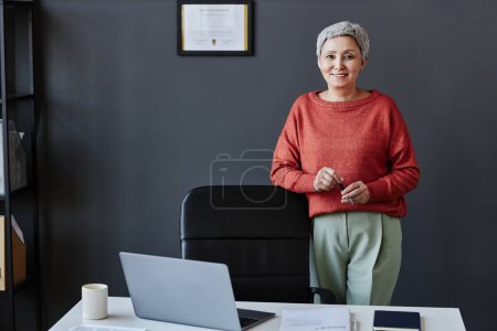 Téléchargez les photos : Portrait d'une aînée réussie debout au bureau, portant une tenue rouge contrastée et souriant à la caméra, espace de copie - en image libre de droit