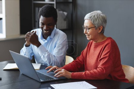Téléchargez les photos : Portrait latéral d'une femme d'affaires âgée souriante utilisant un ordinateur portable avec un collègue au travail au bureau - en image libre de droit