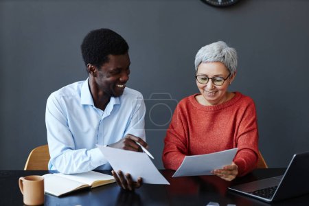 Téléchargez les photos : Portrait d'une femme d'affaires âgée souriante lisant des documents tout en travaillant avec un collègue au bureau - en image libre de droit