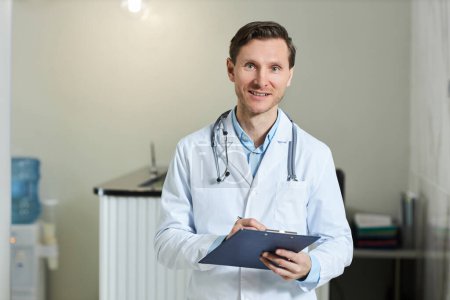 Photo for Waist up portrait of smiling man as doctor wearing lab coat in clinic and looking at camera holding clipboard, copy space - Royalty Free Image