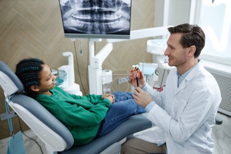 Téléchargez les photos : Dentiste souriant tenant un modèle de dent tout en consultant un enfant patient dans une clinique dentaire, espace de copie - en image libre de droit