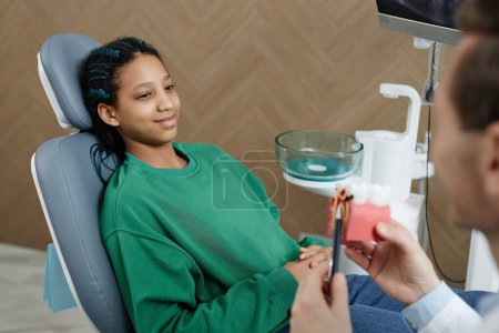 Téléchargez les photos : Portrait de fille noire souriante dans une clinique dentaire assise sur une chaise et écoutant un dentiste - en image libre de droit