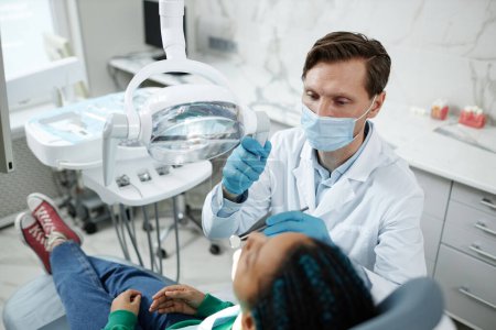 Photo for High angle view at male dentist wearing mask during child checkup in dental clinic - Royalty Free Image