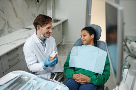 Téléchargez les photos : Portrait grand angle d'une adolescente souriante dans une chaise dentaire écoutant un dentiste lors d'une consultation - en image libre de droit
