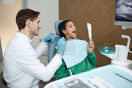 Téléchargez les photos : Vue latérale portrait d'une adolescente noire regardant dans un miroir lors d'un examen dentaire à la clinique - en image libre de droit