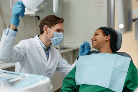 Photo for Side view portrait of friendly male dentist working with teenage girl in dental clinic - Royalty Free Image