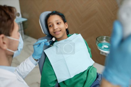 Téléchargez les photos : Portrait à angle élevé d'une adolescente noire assise sur une chaise dentaire lors d'un examen dentaire à la clinique - en image libre de droit