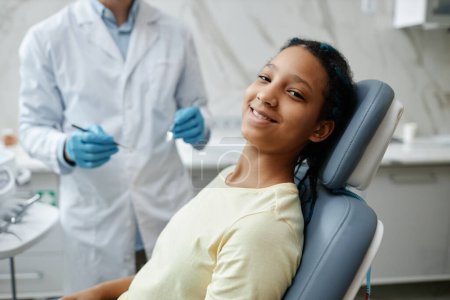 Photo for Portrait of smiling black girl sitting in dental chair during checkup and looking at camera - Royalty Free Image