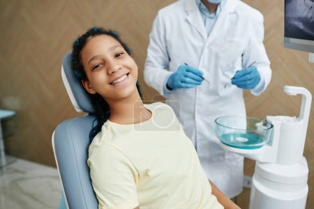 Téléchargez les photos : Portrait grand angle de la jeune fille noire souriante assise dans une chaise dentaire et regardant la caméra pendant le check-up - en image libre de droit