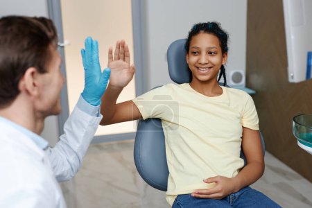 Photo for Portrait of teen black girl high-fiving dentist while enjoying dental care appointment in clinic and smiling - Royalty Free Image