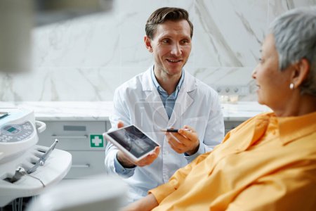 Téléchargez les photos : Portrait d'un dentiste souriant tenant une tablette numérique avec une image radiographique des dents tout en expliquant la chirurgie dentaire au patient - en image libre de droit