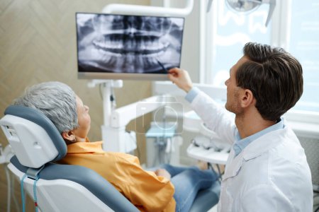 Foto de Vista posterior del dentista masculino apuntando a la imagen de rayos X del diente en la pantalla durante la consulta en la clínica dental moderna - Imagen libre de derechos