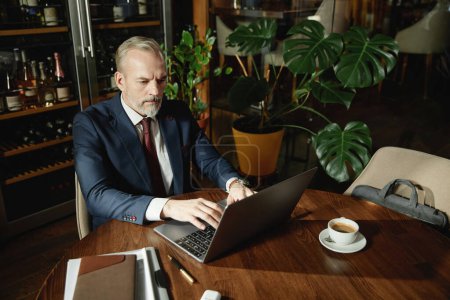 Téléchargez les photos : Homme d'affaires mature se concentrant sur son travail à distance sur ordinateur portable tout en étant assis à table dans un café - en image libre de droit
