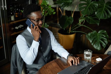 Téléchargez les photos : Homme d'affaires afro-américain parlant sur smartphone et travaillant en ligne sur ordinateur portable tout en étant assis dans un café - en image libre de droit
