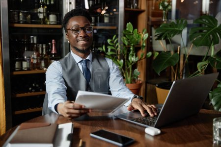 Téléchargez les photos : Portrait de jeune homme d'affaires en lunettes souriant à la caméra tout en travaillant avec un ordinateur portable et un document dans un café - en image libre de droit