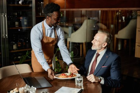 Téléchargez les photos : Serveur afro-américain servant le repas à un homme d'affaires pendant le déjeuner au restaurant - en image libre de droit