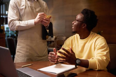 Téléchargez les photos : Homme d'affaires afro-américain faisant l'ordre de servir tout en étant assis à table pendant le déjeuner dans un café - en image libre de droit