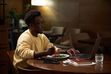 Foto de Hombre de negocios afroamericano que trabaja en línea en el ordenador portátil sentado en la mesa y almorzando en la cafetería - Imagen libre de derechos