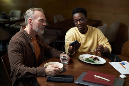 Téléchargez les photos : Partenaires discutant de présentation en ligne sur smartphone pendant le déjeuner d'affaires à table dans un café - en image libre de droit