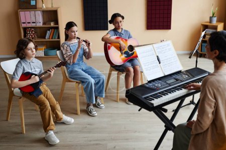 Téléchargez les photos : Groupe d'enfants assis sur des chaises et jouant des instruments de musique avec professeur jouant du piano pendant les cours de musique - en image libre de droit