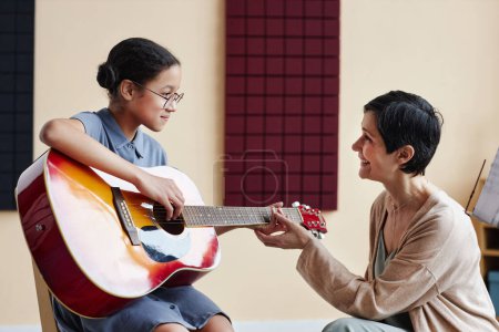 Téléchargez les photos : Professeur souriant enseignant à l'élève à jouer de la guitare pendant les cours de musique à l'école - en image libre de droit