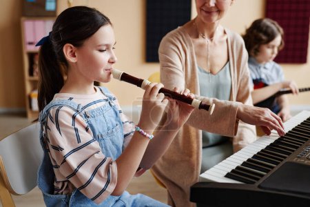 Téléchargez les photos : Écolière jouant de la flûte avec professeur jouant du piano pendant les cours de musique en classe - en image libre de droit