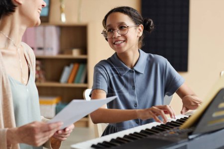 Téléchargez les photos : Fille souriante apprenant à jouer du piano avec professeur tenant des partitions, ils parlent et sourient pendant la leçon de musique - en image libre de droit