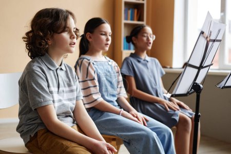 Téléchargez les photos : Groupe d'enfants assis devant une partition sur un stand et chantant ensemble pendant un cours de musique - en image libre de droit