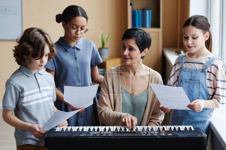 Téléchargez les photos : Professeur mature jouant du piano et répétant la chanson avec les enfants pendant qu'ils se tiennent debout avec des notes autour d'elle en classe de musique - en image libre de droit