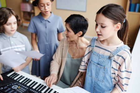 Foto de Coro de niños cantando canciones en clase de música mientras el profesor toca el piano y cantando junto con ellos - Imagen libre de derechos