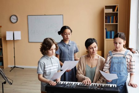 Téléchargez les photos : Groupe d'enfants lisant des notes et chantant des chansons pendant que l'enseignant joue du piano pendant les cours de musique en classe - en image libre de droit